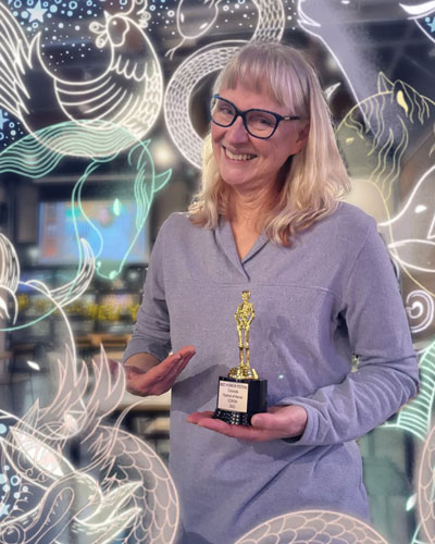 Jeanni, smiling blonde woman with shoulder length hair and black glasses, in a light longsleeve purple shirt, holding an award for Colorado Festival of Horror with an animal filter around the edges